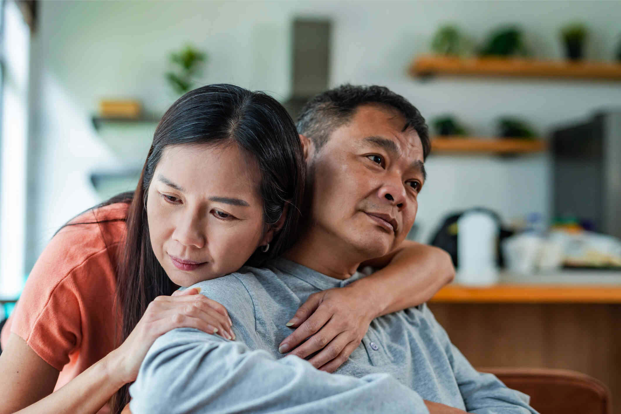 A sad middle aged man sits in his home as his wife stands behind him and wraps her arms around his neck with a worried expression.