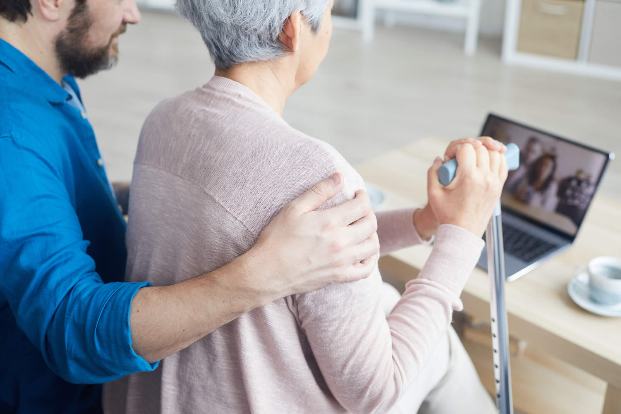 A man and an older woman are sitting down together and looking at a video call on a laptop; the man has his arm around the woman, and she is holding a cane.